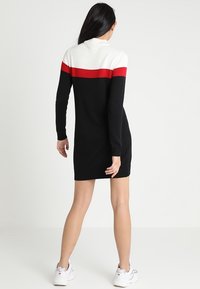 Gebreide jurk met een zwart lichaam en een witte en rode horizontale streep over het bovenste deel. Lange mouwen en een iets getailleerd ontwerp.