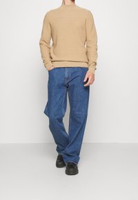 Beige gerippter Pullover kombiniert mit lockeren blauen Denim-Jeans und schwarzen Schnürschuhen. Der Pullover hat einen Rundhalsausschnitt, die Jeans weisen ein klassisches Nähmuster auf.