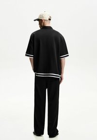 Polo nero a maniche corte con strisce bianche ai bordi delle maniche e sul fondo, abbinato a pantaloni neri larghi. Un cappellino chiaro è posizionato sulla testa.