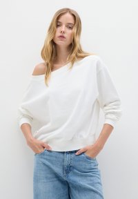 Donna bionda con capelli sciolti indossa una felpa bianca oversize a spalle scoperte e jeans a vita alta blu chiaro, con le mani in tasca.