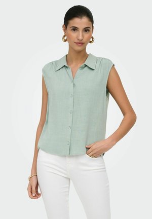 Vrouw met donker haar die gouden oorringen draagt, een mouwloos lichtgroen overhemd met knopen en witte broek, poserend met één hand in de zak.