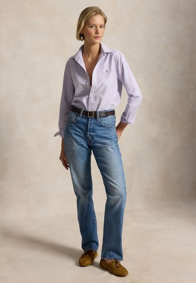Young woman standing, wearing a light purple button-up shirt, blue jeans with a black belt, and brown moccasin shoes, neutral background.