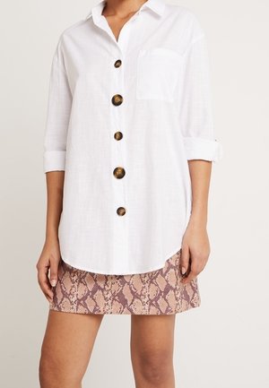 Frau trägt ein weißes, langärmliges Hemd mit Knöpfen und einer Brusttasche sowie einen kurzen braunen Rock mit Schlangenprint.