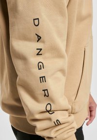 Beige pulóver fekete "DANGEROUS" felirattal függőlegesen a bal ujján, puha tapintással és elülső zsebbel.