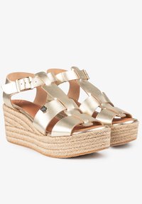 Sandalias cuñas metálicas doradas con tiras entrelazadas, puntera cuadrada y plataforma envuelta en yute. Cuenta con correas en el tobillo ajustables y un acabado texturizado.