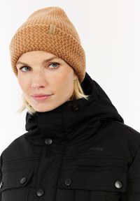 Vrouw met een lichte huid en blauwe ogen, die een bruine gebreide muts en een zwarte winterjas met capuchon en knoopzakken draagt.