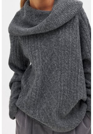 Kvinde iført en grå tekstureret strikket sweater med bred, foldet krave og lange ærmer, kombineret med grå bukser.