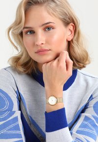 Montre en or avec un cadran rond et un bracelet en maille, dotée d'un cadran blanc et de marqueurs d'heure simples. Le modèle porte un pull à motifs bleus et gris.
