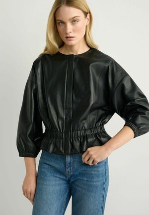 Blondynka w czarnej skórzanej kurtce z krótkim krojem, z elastycznym pasem i rękawami, w połączeniu z niebieskimi, wysokim stanem jeansami, na jednolitym tle.