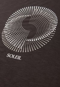 Motivo circolare di punti bianchi che si irradiano su tessuto nero con la parola "SOLEIL" stampata sotto il disegno.