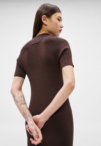 Νεαρή γυναίκα με μακριά σκούρα μαλλιά φοράει ένα εφαρμοστό, ριγωτό, καφέ φόρεμα με κοντά μανίκια, δείχνοντας τα τατουάζ στο αριστερό της πήχη, με τα χέρια της ενώμενα πίσω από την πλάτη.