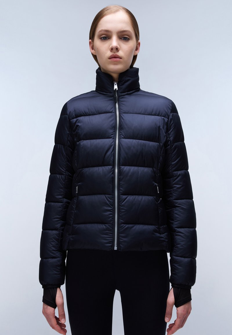 Giacca a vento blu scuro con collo alto, chiusura con zip e design squadrato. Presenta sezioni imbottite e polsini in maglia estesi con fori per il pollice.