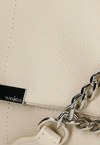 Crème leren handtas met een gestructureerde afwerking, zilveren kettingriem en een zwart metalen logo accent. Compacte, slanke rechthoekige vorm.