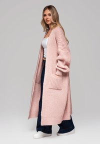 Dlouhá, růžová pletená kardigan s volným střihem, ribbovanou texturou, bočními kapsami a otevřeným předním dílem, stylizovaná přes bílé tričko a tmavé džíny.