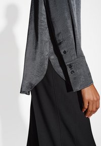 Camicia grigia testurizzata con orlo curvo, dotata di polsini con bottone e tessuto liscio, abbinata a un indumento nero sullo sfondo.