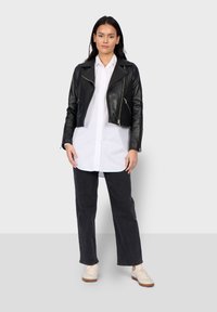 Černá kožená motorkářská bunda s vykrojeným designem, zipy a širokými klopami, nošená přes bílé tričko a tmavé kalhoty. Světlé tenisky.