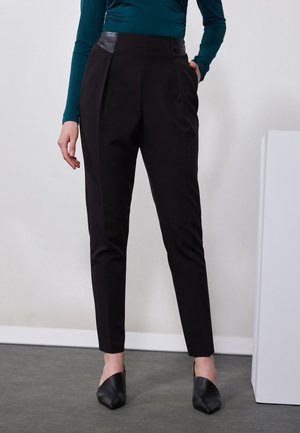 Black tailored trousers featuring a smooth finish, pleat details, and leather accents at the waistband. Paired with black pointed-toe shoes.