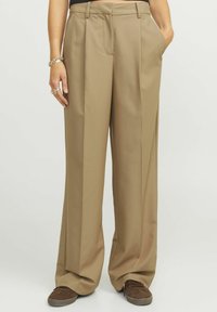 Pantalons larges marron clair avec un devant plissé, des poches latérales et une texture lisse, assortis à des baskets marron foncé.