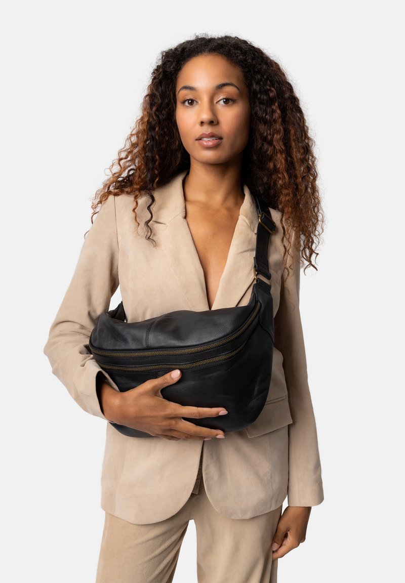 Borsa a tracolla in pelle nera con design curvato, cerniere in tonalità oro e tracolla regolabile, appoggiata contro un blazer color avorio.