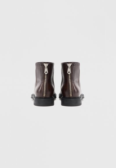 Um par de botas de tornozelo em couro castanho escuro com fechos zip em prata na parte de trás e solas pretas grossas, sobre um fundo branco.