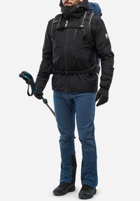 Schwarze isolierte Jacke, blaue technische Hose, Handschuhe, Wanderstöcke und Schuhe. Rucksack mit blauen Akzenten und mehreren sichtbaren Riemen.