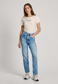 Camiseta blanca con rayas amarillas y logo negro, combinada con jeans de tiro alto azul claro y zapatillas gris y blancas.