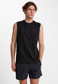 Svart ärmlös bomullströja med rund halsringning, prydd med en liten röd chevron-logotyp på vänster bröst. Slät struktur, avslappnad passform.