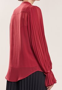 Rød, gennemsigtig bluse med lodrette plisseringer på lange ærmer og en bundet manchet, der har en rund kant og et enkelt kravedesign.