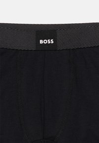 Μαύρο ανδρικό εσώρουχο από ύφασμα με εμφανείς ραφές και γκρι ζώνη μέσης που φέρει το λευκό λογότυπο της μάρκας BOSS στο κέντρο.