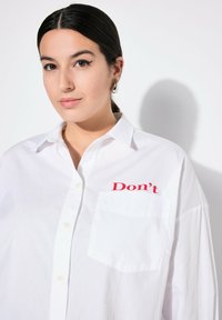 Camicia bianca con bottoni, caratterizzata da un testo ricamato rosso "Don't" sulla tasca sinistra del petto, con colletto classico e design dalla vestibilità rilassata.