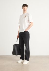 Maglietta bianca a maniche corte con logo, jeans neri dritti e sneakers bianche. Tiene una borsa tote nera. Tessuto di cotone liscio.
