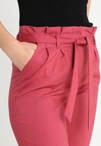 Pantalons taille haute de couleur rose avec une large ceinture, poches latérales et plis. Fabriqués en tissu lisse, présentant un design ajusté pour un look poli.