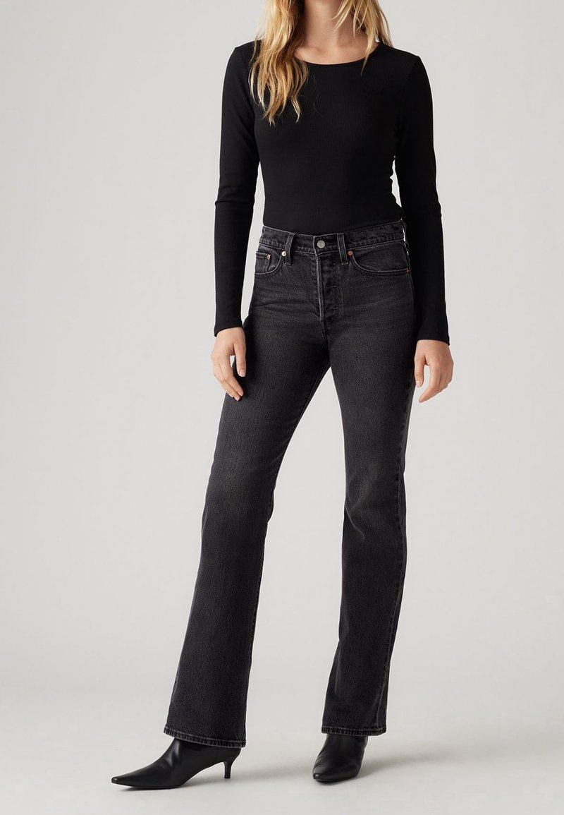 Femme portant un haut noir à manches longues, un jean taille haute gris foncé et des bottines noires à bout pointu, debout sur un fond uni.