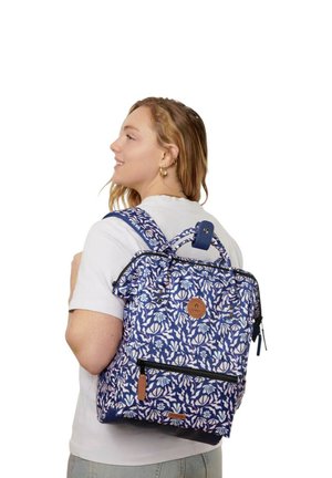 Woman wearing blue and white floral patterned backpack with front zipper pocket and top handle, facing left, smiling.