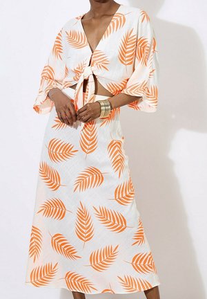 Femme portant une tenue deux pièces blanche avec un motif de feuilles orange, un top noué à l'avant et une jupe assortie, accessoirisée de bracelets dorés.
