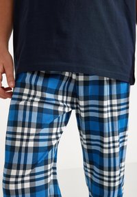 Pantalon de pyjama à carreaux bleus et noirs avec des accents blancs ; fabriqué en tissu de coton doux. Le motif présente des carrés et des rayures alternés.