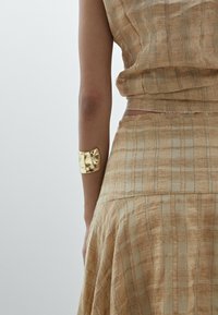 Beiges Zweiteiler-Outfit mit einem strukturierten, gewebten Muster und goldenem, metallic glänzendem Armreif, das eine lässige, figurbetonte Silhouette bietet.