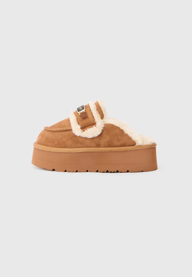 Tan slip-on tossut, joissa on shearling-vuori, tasapohjainen pohja, ommeltu muotoilu ja säädettävä hihna metalliosilla.
