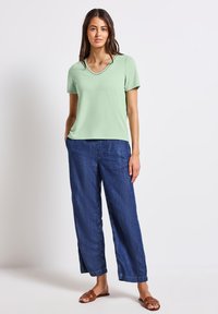 Zielona koszulka z krótkim rękawem i dekoltem w serek, ozdobiona haftem, zestawiona z luźnymi ciemnoniebieskimi dżinsami oraz brązowymi płaskimi sandałami.