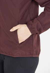 Bordeaux lehká bunda s kapsou na zip, elastickými manžetami a texturovaným povrchem, s uvolněným střihem pro pohodlí a pohyblivost.
