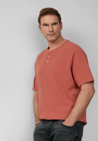 Koszulka z krótkim rękawem w kolorze koralowym o fakturowanym wykończeniu, z zapięciem henley na guziki. Noszona z ciemnoszarymi dżinsami, ręce w kieszeniach.