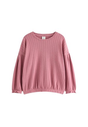 Junges Mädchen mit geflochtenen Haaren, das einen rosa gerippten Pullover und passende Hose trägt, lächelt und steht mit leicht ausgestreckten Armen.