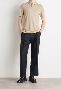 Beige polo shirt with three gray buttons and a subtle logo, paired with black trousers and black loafers, standing on a neutral floor.