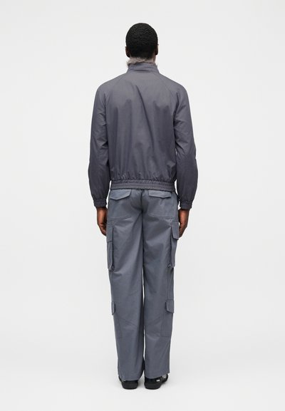 Γκρί νάιλον μπουφάν με όρθιο γιακά και ελαστική μέση, συνδυασμένο με γκρί παντελόνι καρό με πολλές τσέπες και ίσια μπατζάκια.