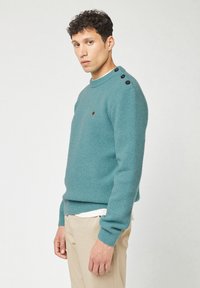 Mann in einem strukturierten türkisfarbenen Pullover mit drei schwarzen Knöpfen an der Schulter, beigen Hosen, der schräg vor einem einfachen, hellen Hintergrund steht.