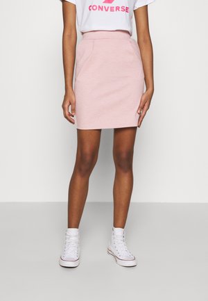 Kvinde iført en lyserød nederdel over knæet, hvid Converse t-shirt og hvide høje sneakers, stående foran en ensfarvet baggrund.