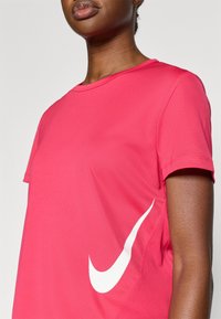 Ροζ μπλουζάκι Nike με κοντά μανίκια και στρογγυλή λαιμόκοψη· διαθέτει μεγάλο λευκό λογότυπο στην πλ�ευρά και υφή υφάσματος λεία και ελαφριά.