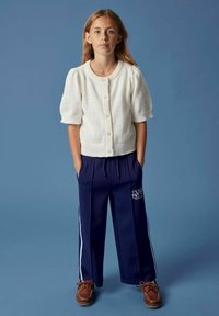 Jeune fille se tenant devant un fond bleu, portant un cardigan blanc à manches courtes et boutonné, un pantalon large bleu marine avec des rayures blanches et des chaussures marron.