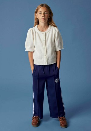 Junges Mädchen steht vor blauem Hintergrund, trägt eine weiße kurzärmlige Knopfleiste-Cardigan, navyfarbene weit geschnittene Hosen mit weißen Streifen und braune Schuhe.