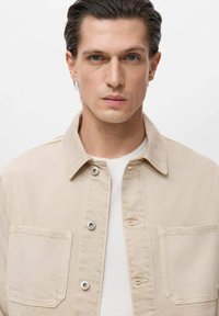 Beige denimjack met een kraag, knoopsluiting en twee borstzakken. Draagt over een wit T-shirt met ronde hals.